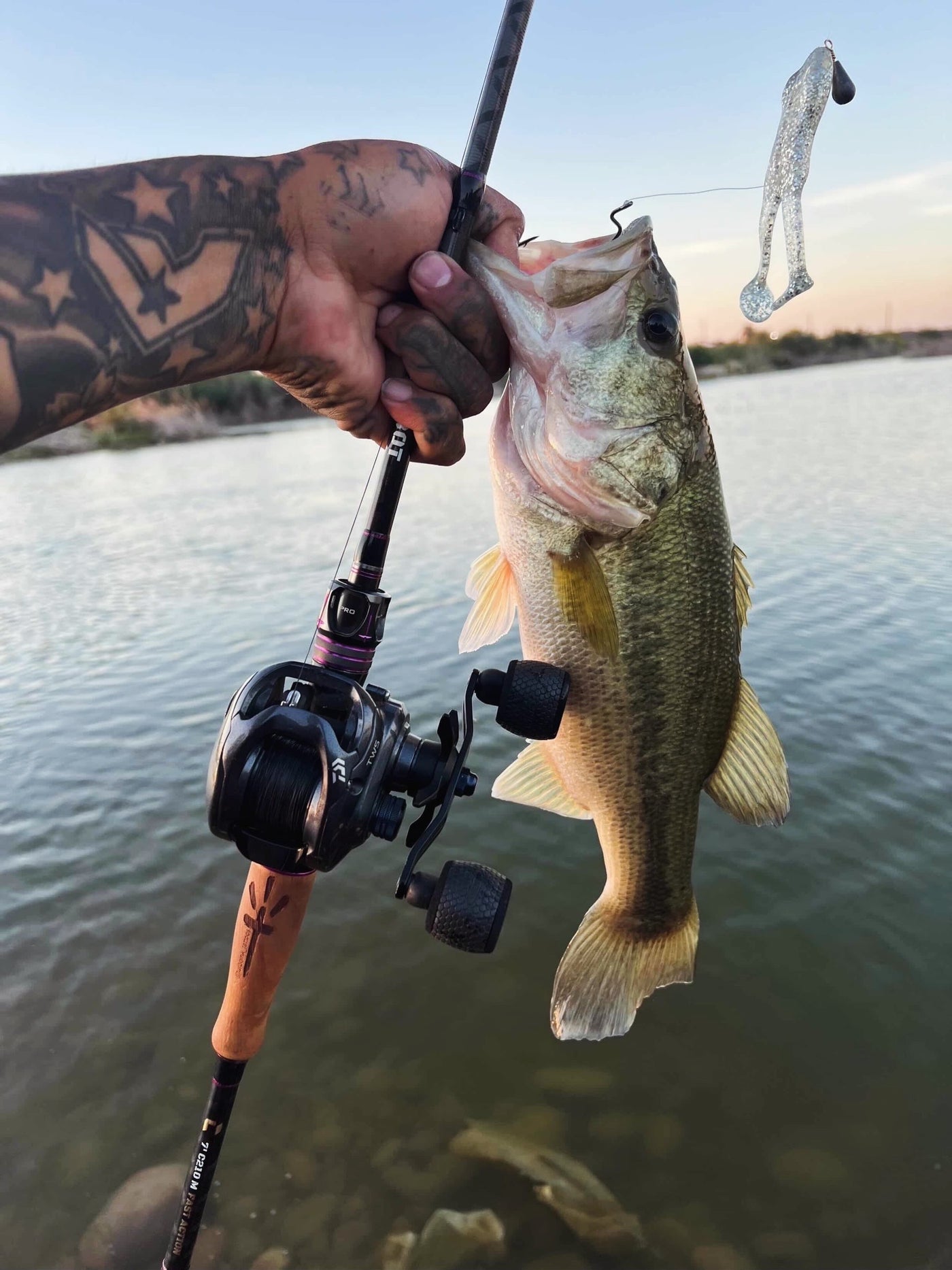Angler holding a bass with a fishing rod featuring a wood handle by a lake at sunset.