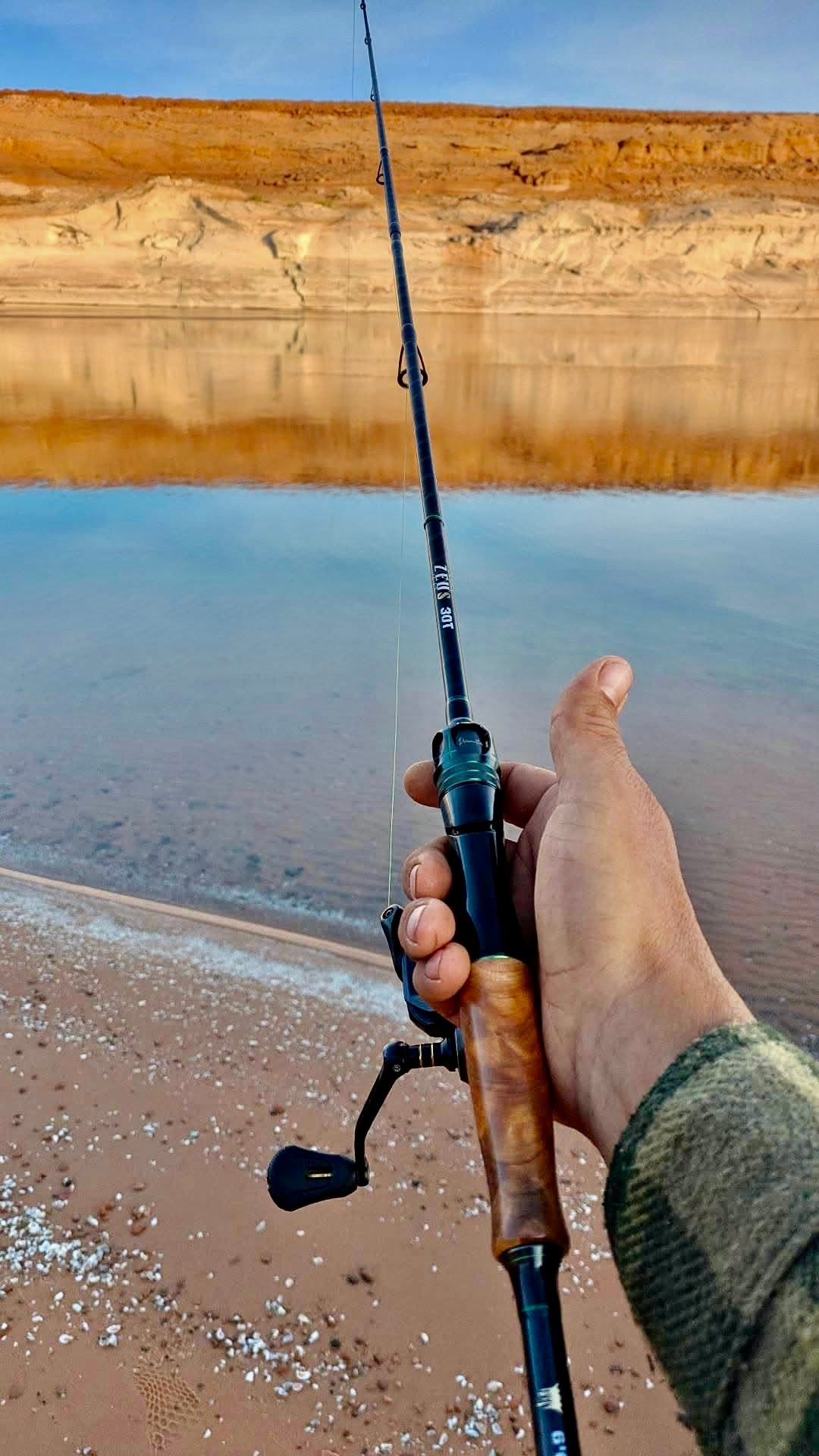 Hand holding a fishing rod with wood handle by a calm lake and sandy shore