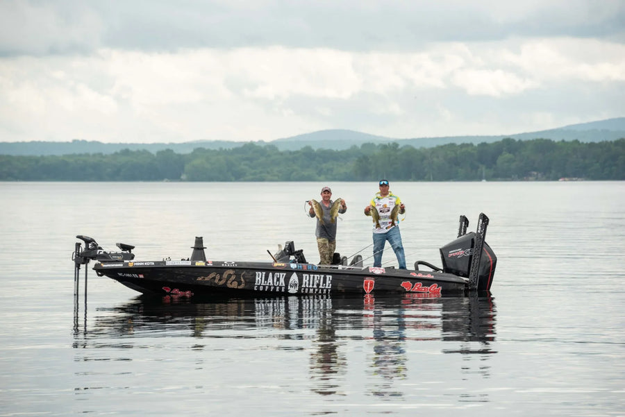 Two anglers on a fishing boat holding up large fish on a calm lake with forested hills