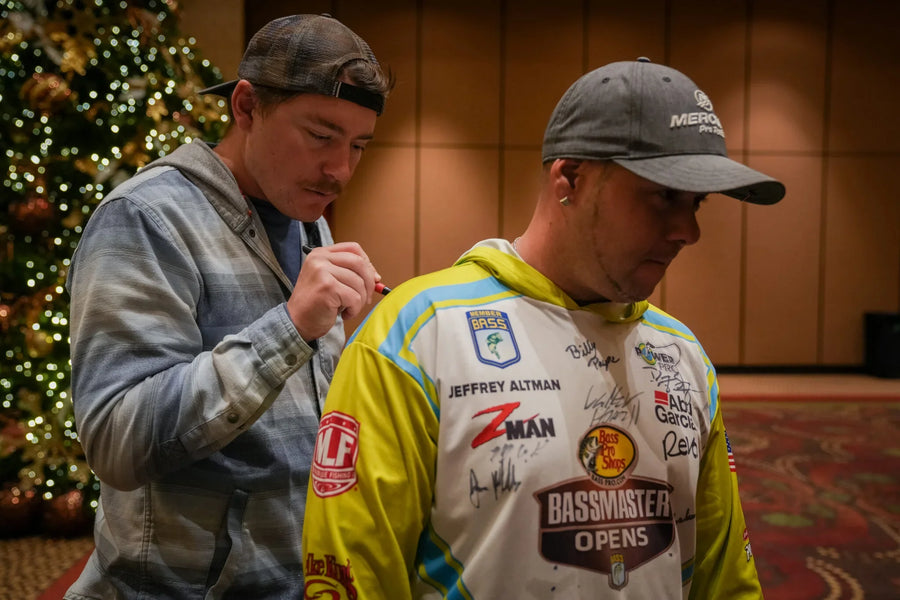 Man signing a fishing jersey at indoor event, Christmas tree in background, sports patches visible