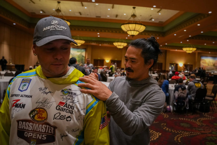 Man signing fishing tournament jersey in indoor event hall, surrounded by attendees.