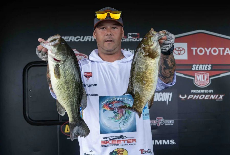 Angler in tournament gear holding two large bass fish at a fishing competition stage